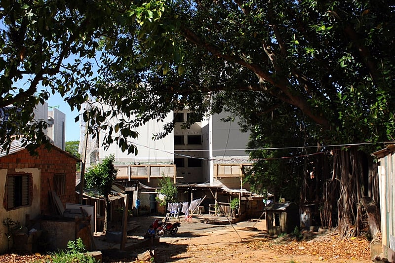 A imagem mostra uma algumas casas em um terreno, cobertas pela sombra de uma grande árvore, ao fundo a construção de um prédio em concreto.